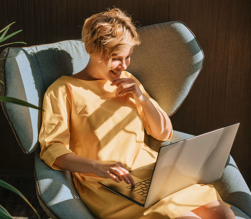 A smiling woman in a yellow dress uses a laptop while sitting in a blue armchair.