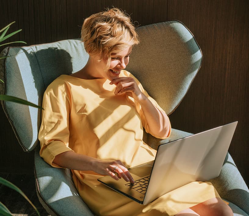 Eine lächelnde Frau in einem yellow Kleid benutzt einen Laptop, während sie in einem blauen Sessel sitzt.