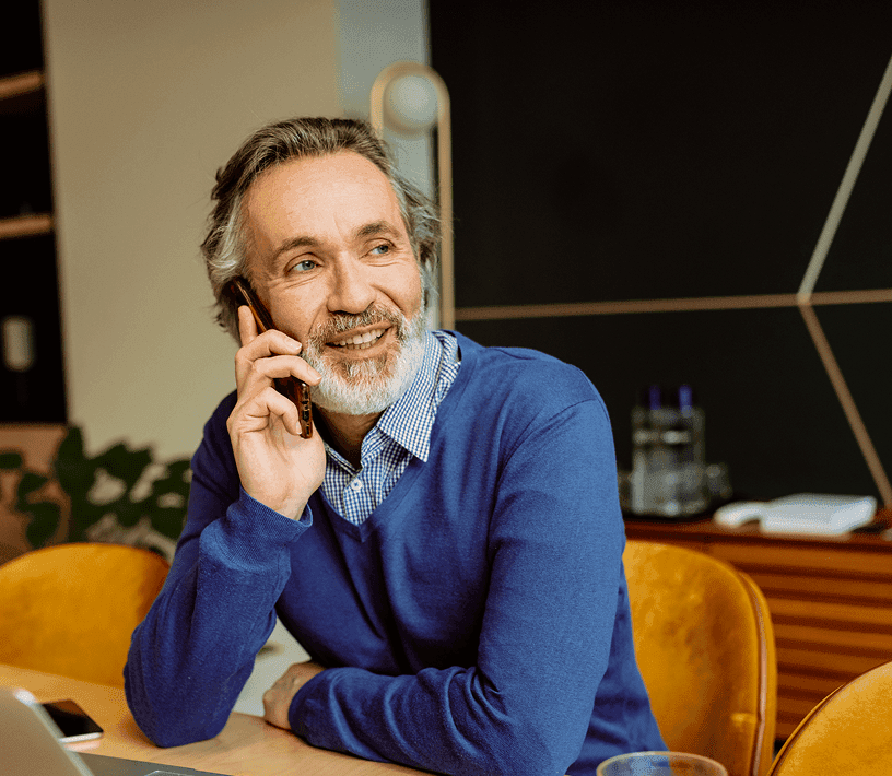 Ein lächelnder, bärtiger Mann in einem blauen Pullover spricht auf einem Smartphone.